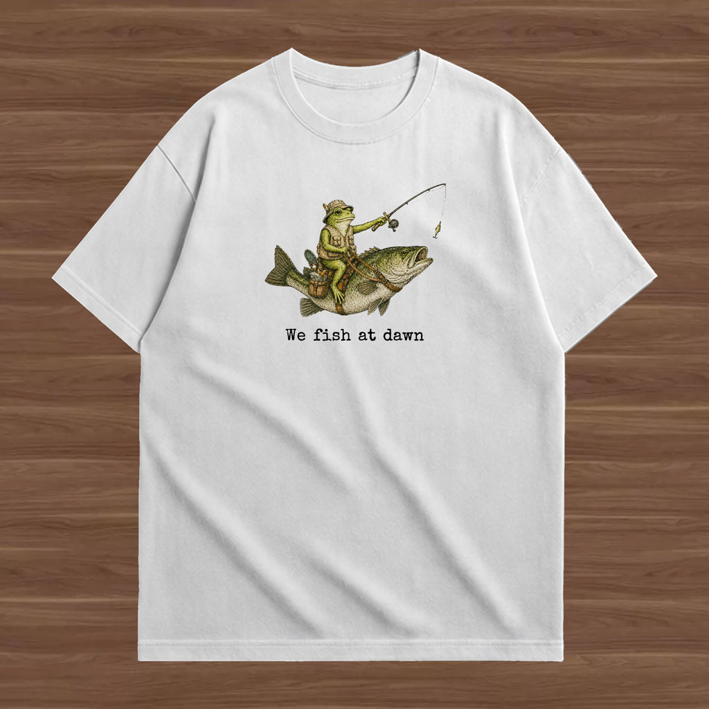 A white short-sleeve crewneck t-shirt laid flat on a brown wood-grain background. The front of the shirt features a graphic of a person in a yellow life vest and green pants fishing on a large fish, with the text 'We fish at dawn' printed below in black.