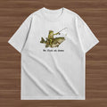 A white short-sleeve crewneck t-shirt laid flat on a brown wood-grain background. The front of the shirt features a graphic of a person in a yellow life vest and green pants fishing on a large fish, with the text 'We fish at dawn' printed below in black.