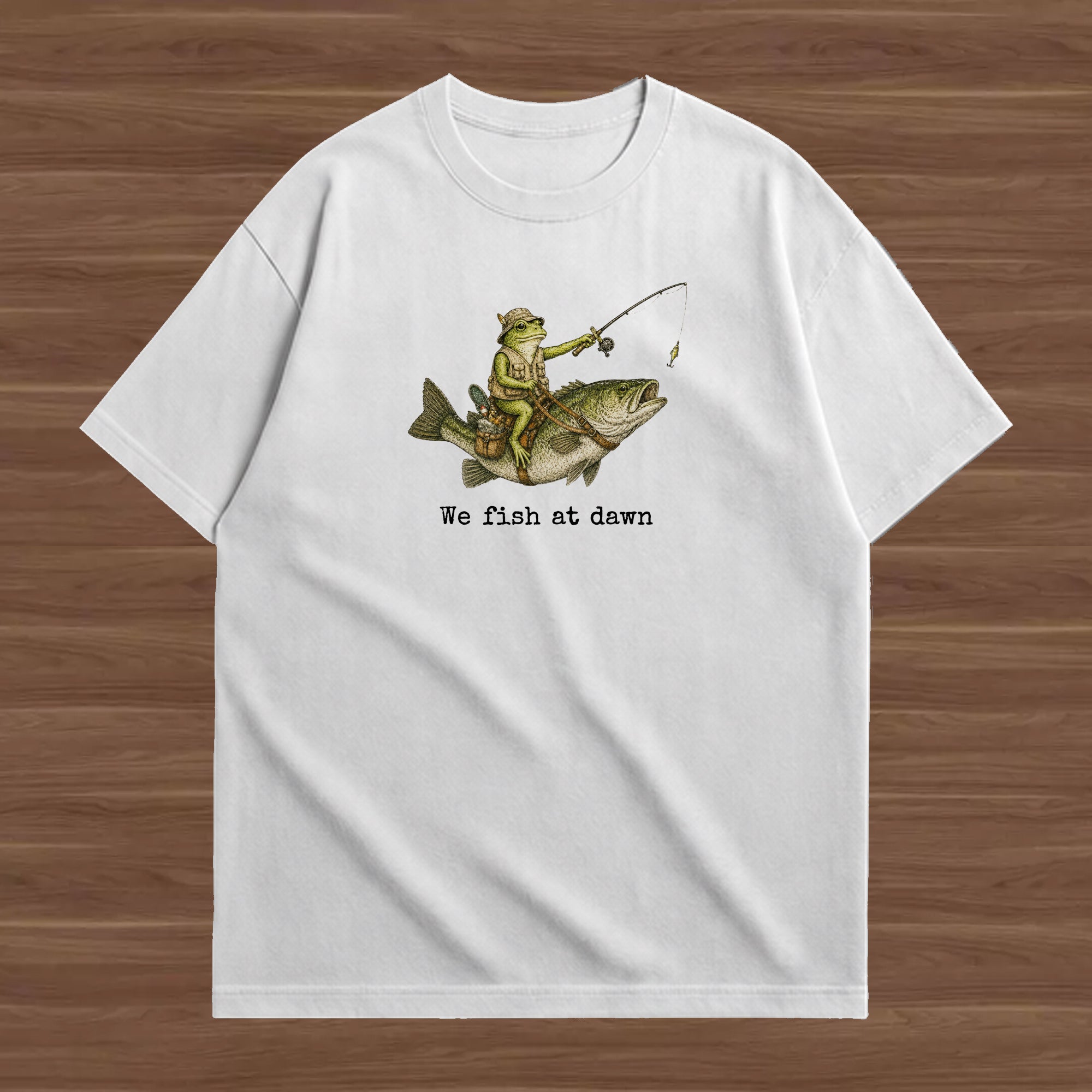 A white short-sleeve crewneck t-shirt laid flat on a brown wood-grain background. The front of the shirt features a graphic of a person in a yellow life vest and green pants fishing on a large fish, with the text 'We fish at dawn' printed below in black.